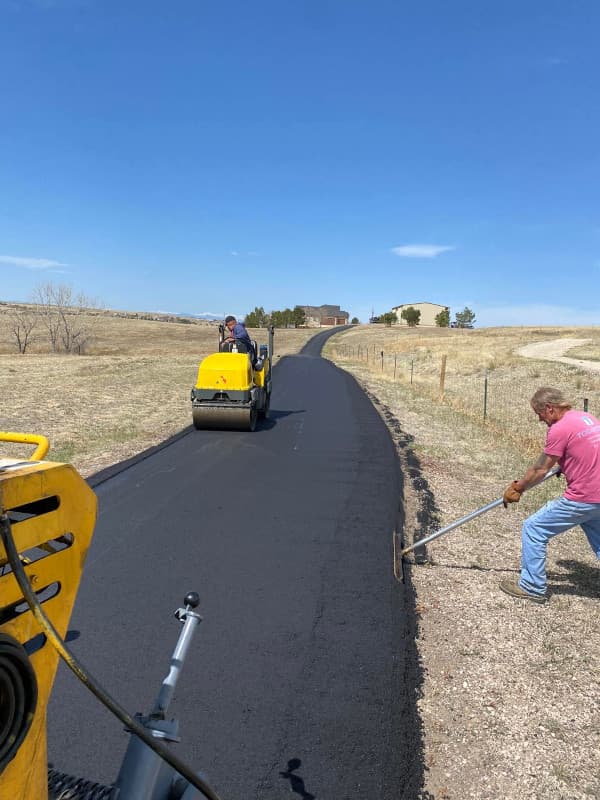 Workers laying asphalt on a rural driveway with a paver under a clear sky.