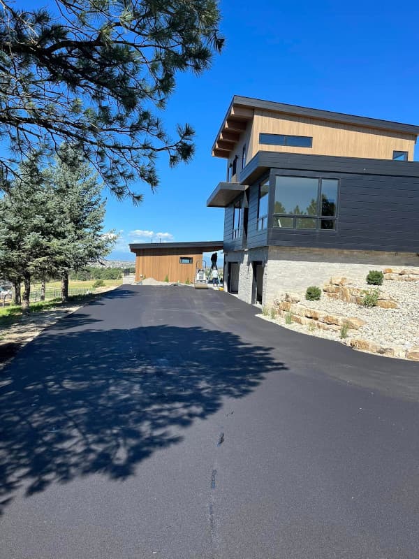 A freshly paved asphalt driveway leading to a modern house with stone and dark siding, set against a clear blue sky with pine trees